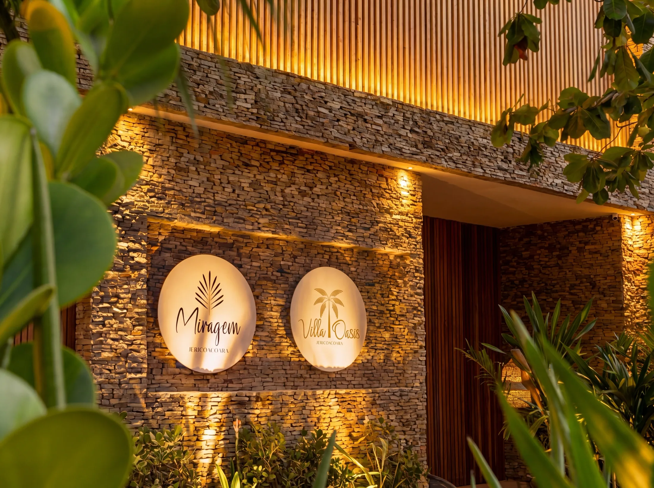 Os letreiros do Miragem e da Villa Oasis iluminados lado a lado na mesma parede de pedra ao entardecer.